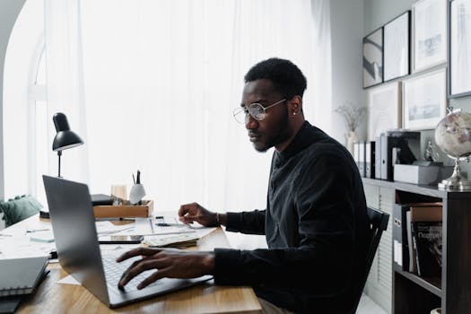pexels-photo-6694489-6694489 Black man in glasses working on laptop at office desk, surrounded by documents and modern decor.